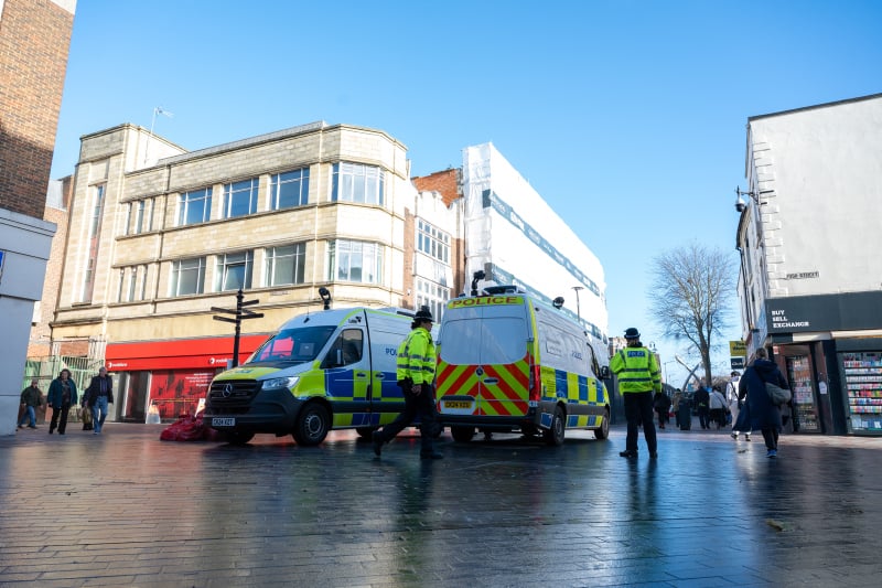 Two police vans equipped with live facial recognition technology are parked in a town centre in from of a large white building. There are two police officers in high visibility yellow jackets walking in front of the vans.